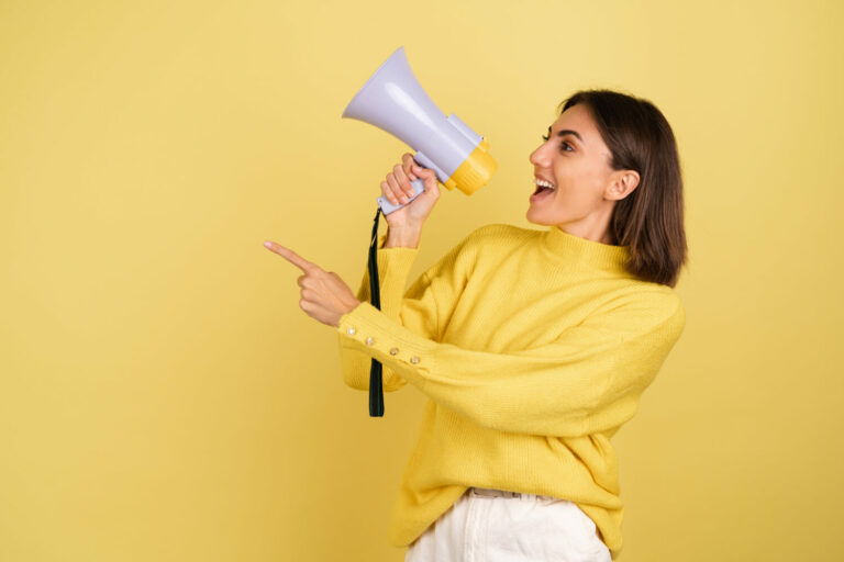 Young woman in yellow warm sweater with megaphone speaker screaming to the left point index finger happy excited