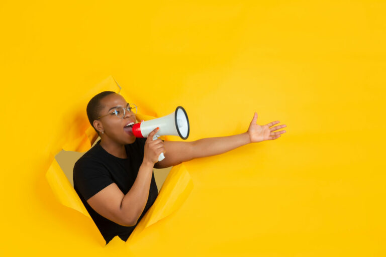 Cheerful young woman poses in torn yellow paper hole background, emotional and expressive, shouting and calling with speaker
